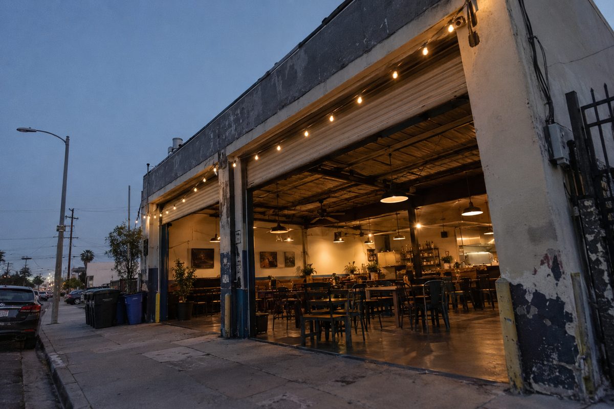 Photo illustration: A former auto-repair garage converted into a neighborhood restaurant on a low-rise Los Angeles street, seen from the sidewalk across the street at dusk, its roll-up garage doors raised open, string lights hung across the front, tables and chairs visible inside under the room's own lamps, the sky fading to deep blue overhead; no people, and no readable lettering, signs or logos.