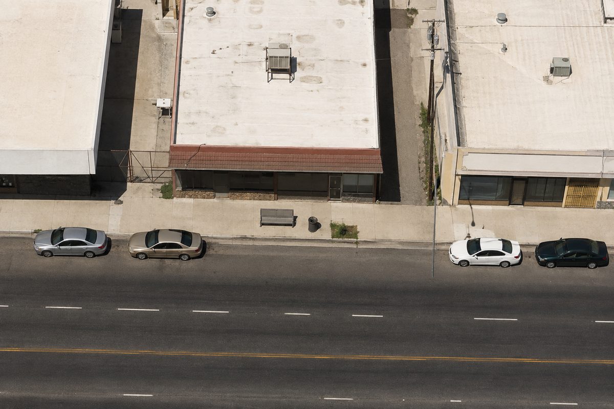 Overhead view at midday of a low commercial strip: stucco storefronts, flat roofs, chain-link fencing, a bus bench and parked cars along the curb.