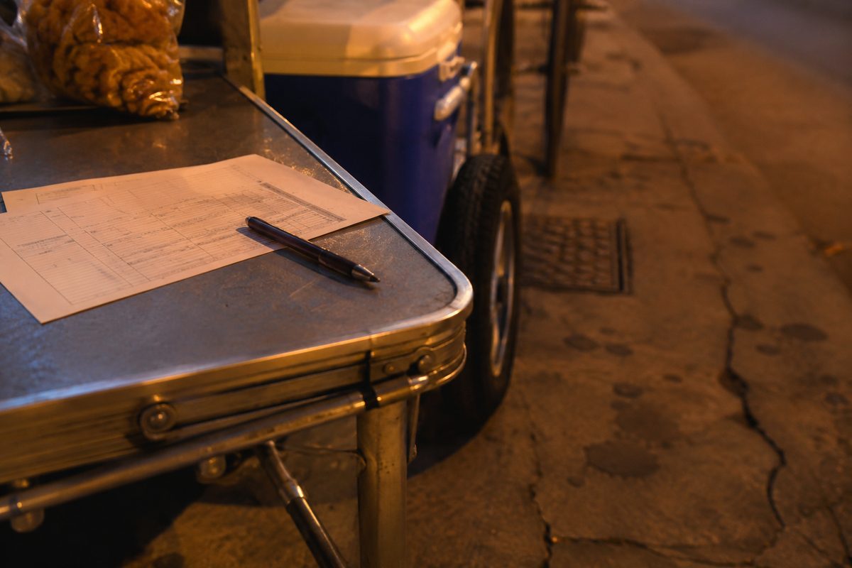 Close-up of a street vendor's folding table and cart wheels on a city sidewalk at dusk, with a paper citation form resting on the table under orange streetlight.