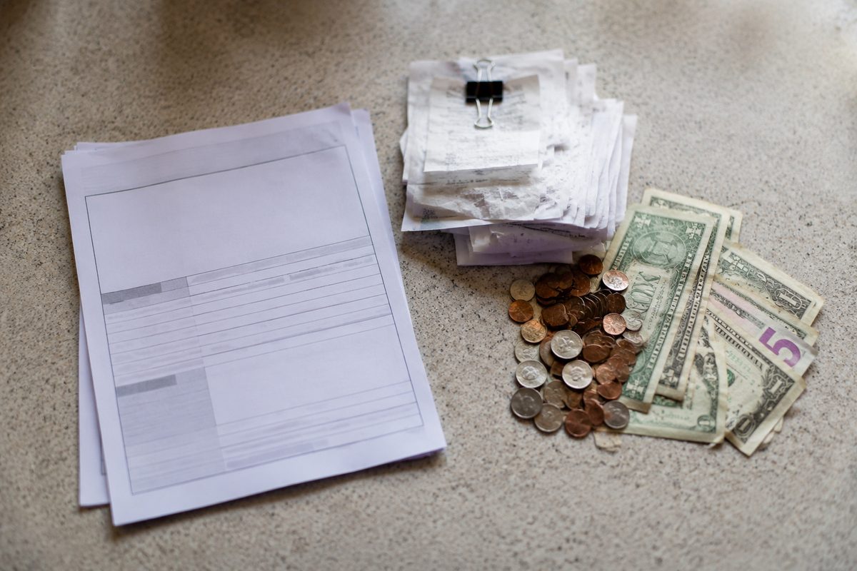 Photo illustration: overhead flat-lay of a plain court filing packet on a counter — a claim form, a stack of receipts held by a clip, and a small pile of coins and bills beside it, the money pile larger than the form; flat neutral daylight, no people, no faces, no courthouse signage, no seals or agency marks.