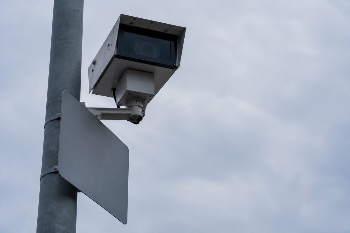 Photo illustration: stylized photographic still life, muted daylight — an unbranded roadside pole-mounted enforcement camera housing seen from below against flat sky, a blank rectangular sign plate beside it, no readable type, no plates, no people, no named streets