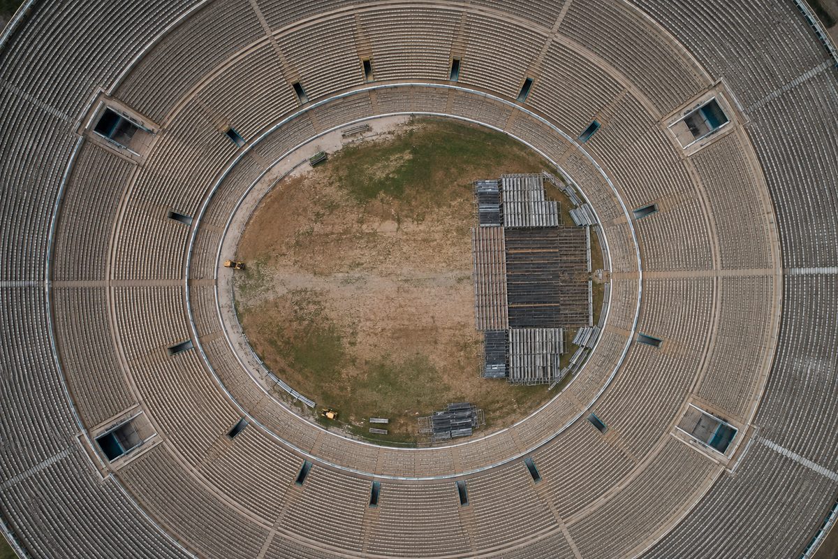 Overhead view of an empty stadium bowl at dawn, concentric rows of seats around a bare field, with stacked scaffolding at one end.