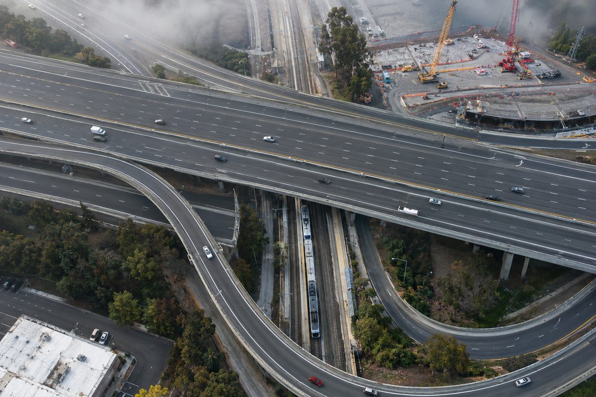 Overhead view of a freeway interchange and a rail line in hazy morning light, with construction cranes in the distance.