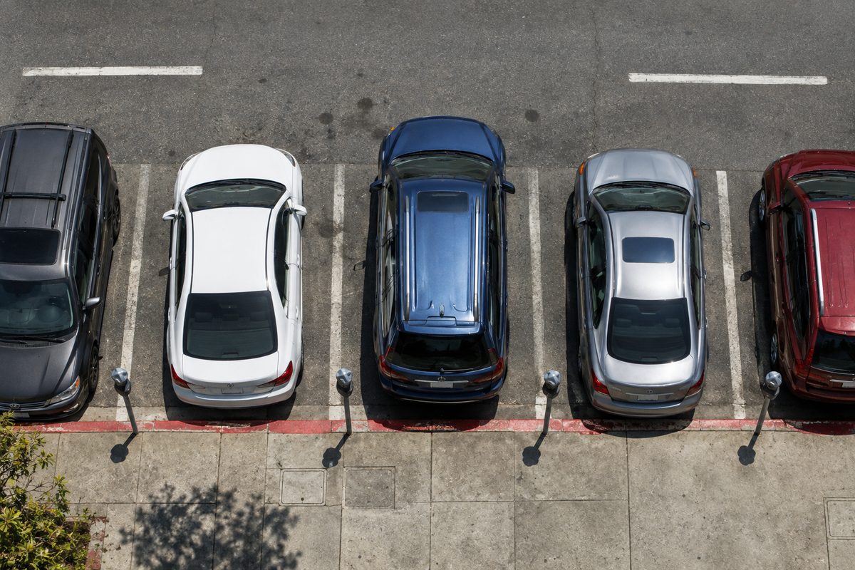 Overhead view of cars parked along a metered curb at midday, shadows short and hard.