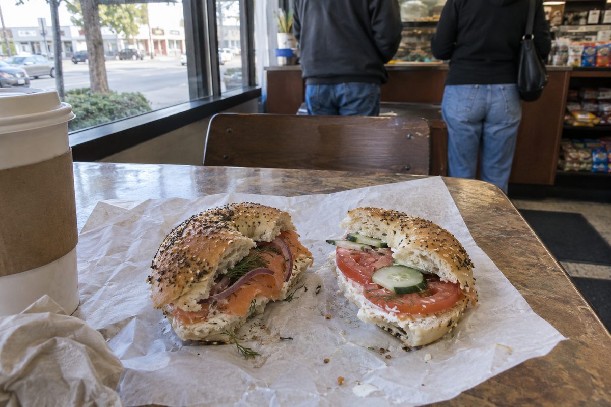 Two halves of bagel sandwiches with smoked salmon, tomato and dill on paper at a deli table by a front window in Highland Park, Los Angeles, with a coffee cup beside them.