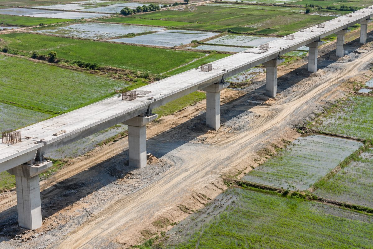 Overhead view at midday of an unfinished concrete rail viaduct crossing flat farmland, with columns, rebar and a gravel haul road below.