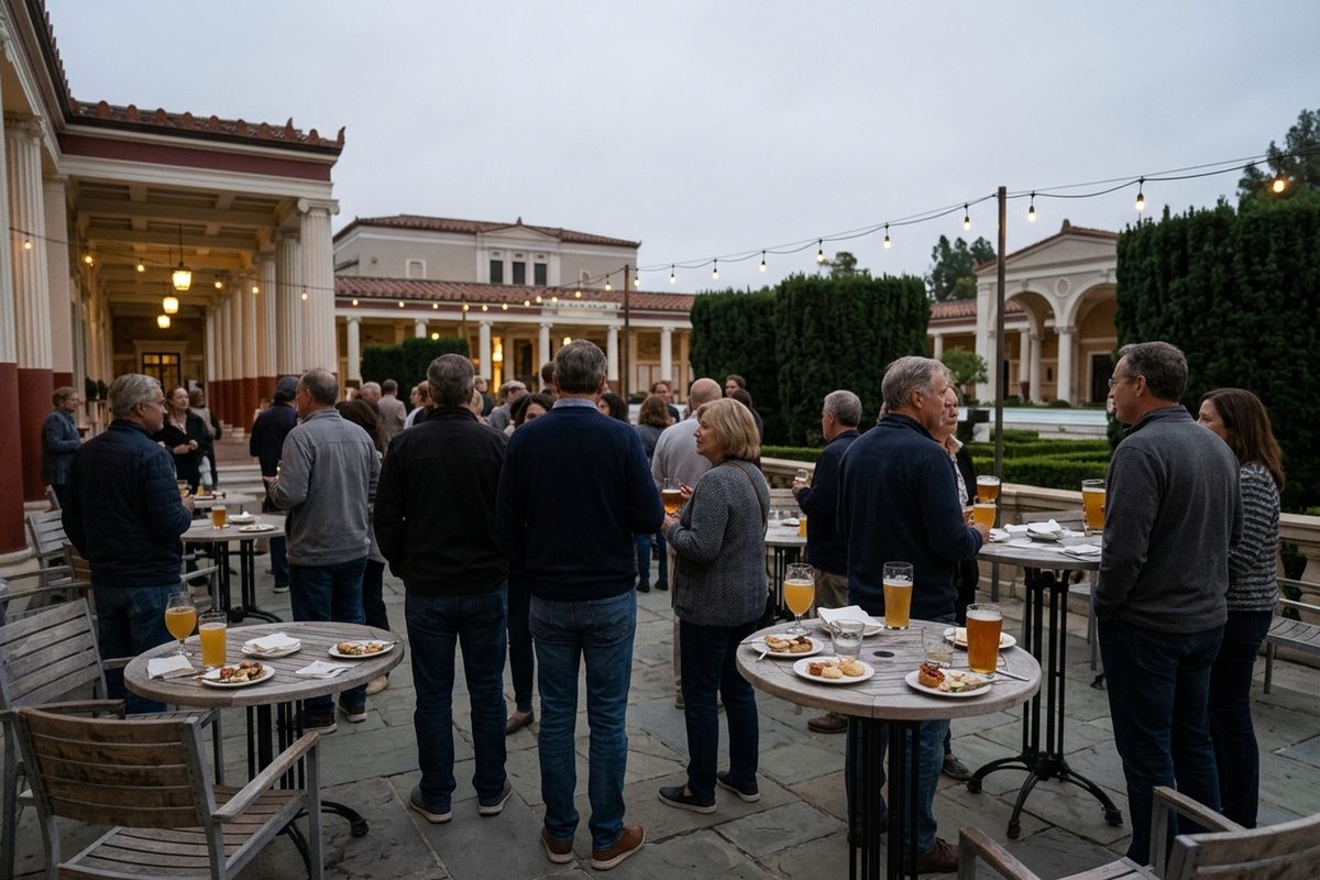 Guests holding glasses of cloudy and amber beer stand among outdoor tables on a museum café terrace in Pacific Palisades under an overcast evening sky.