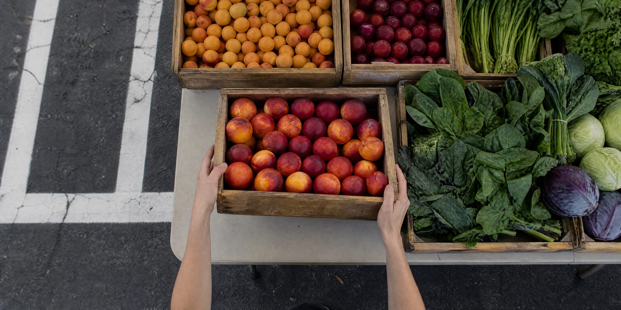 Overhead view of a folding market table with wooden crates of stone fruit and leafy greens, two hands squaring a crate, asphalt and painted parking-stall lines at the table's edge.