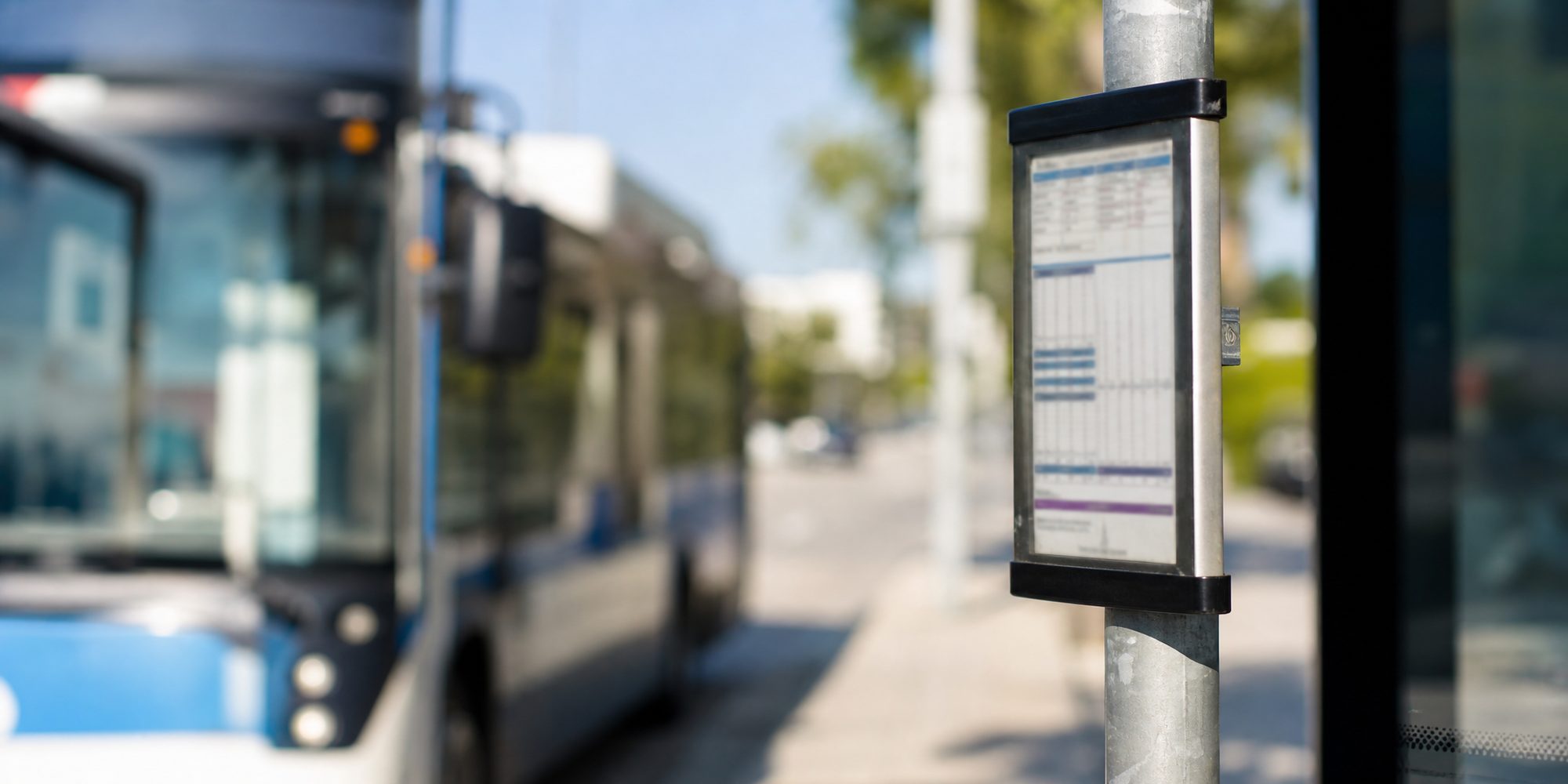 Close-up of a metal bus stop pole with a posted schedule placard, type unreadable, a bus mirror blurred behind it in midday sun.