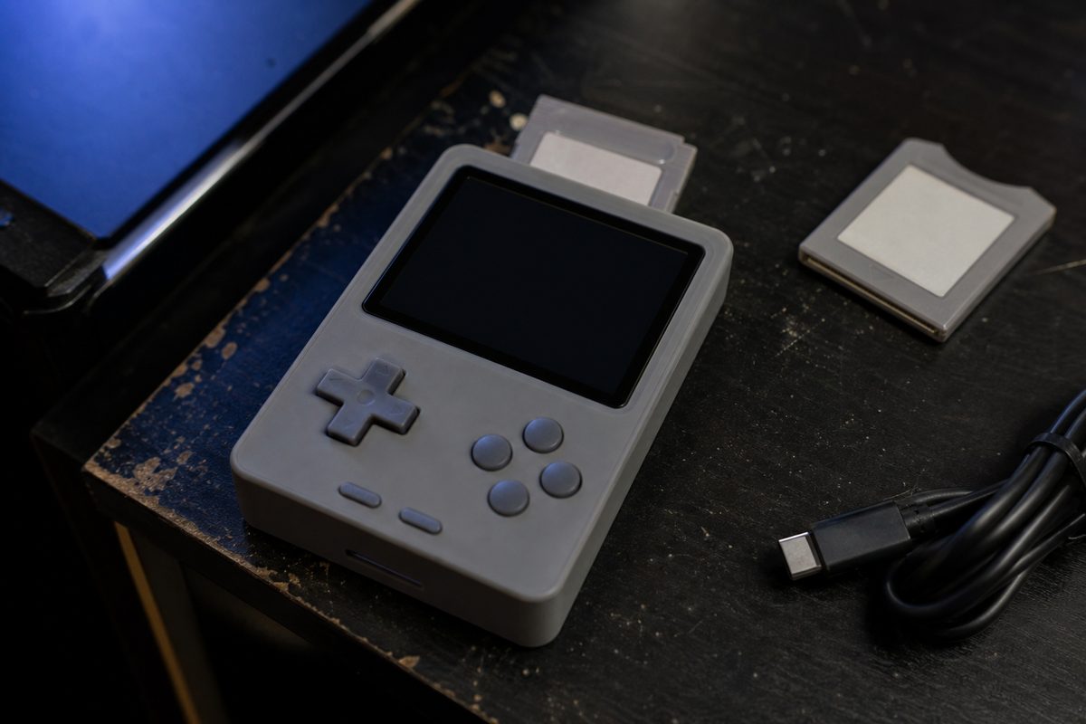 Close macro of a small unbranded handheld game console on a desk beside two blank-labeled cartridges, lit by monitor glow.