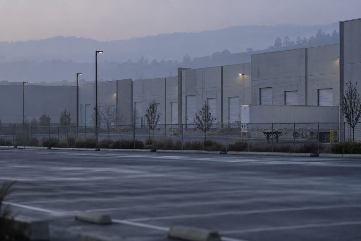 A row of windowless tilt-up industrial buildings behind chain-link fence at dawn, seen compressed by a long lens across an empty parking lot.