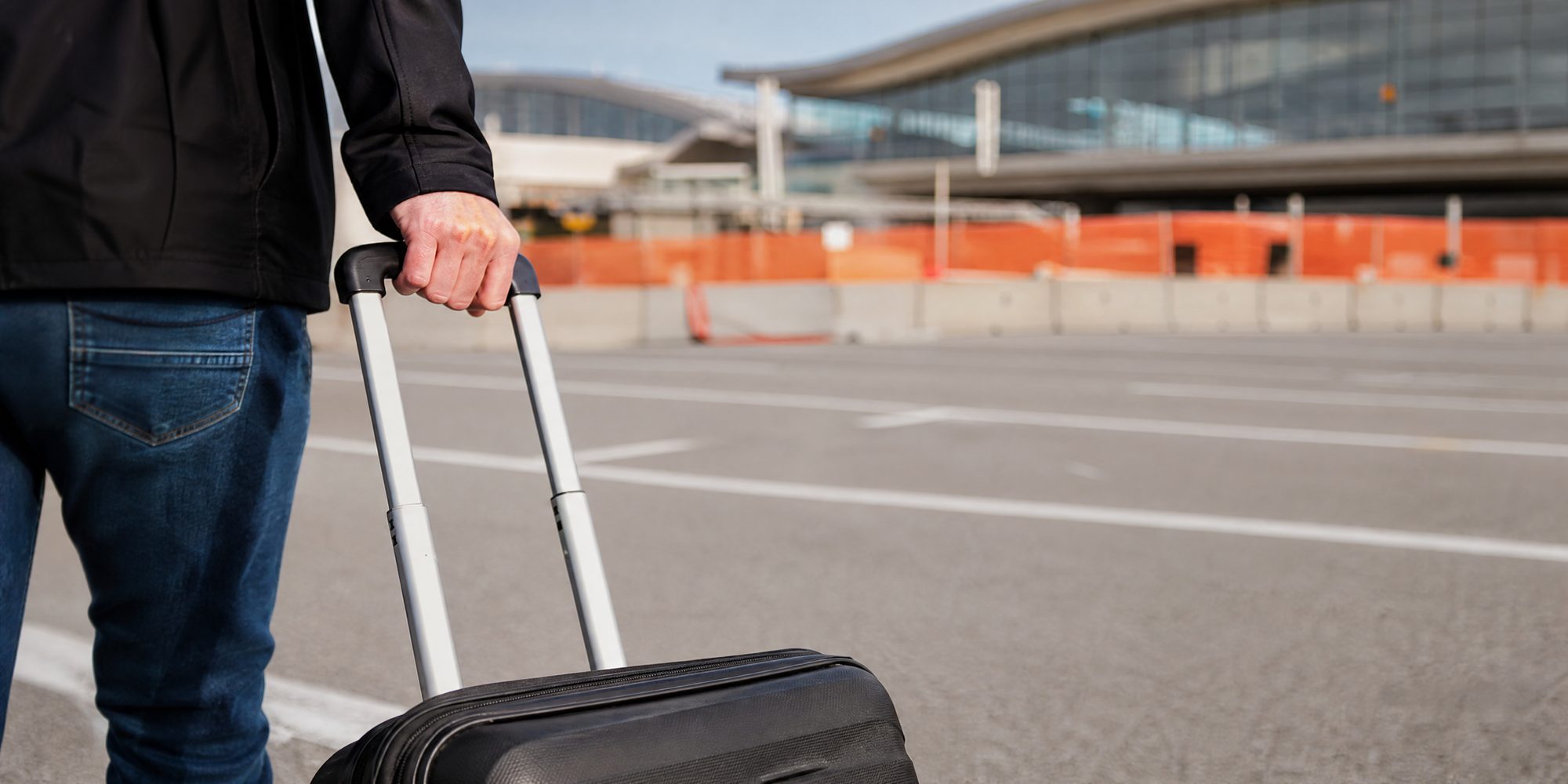 Close-up of a traveler's hand gripping a rolling suitcase handle on sun-bleached asphalt outside an airport terminal, orange construction fencing behind.
