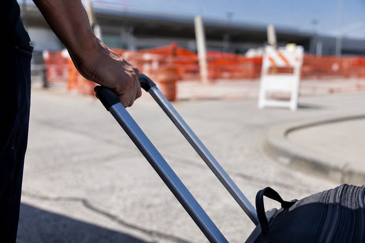 Close-up of a traveler's hand gripping a rolling suitcase handle on sun-bleached asphalt outside an airport terminal, orange construction fencing behind.