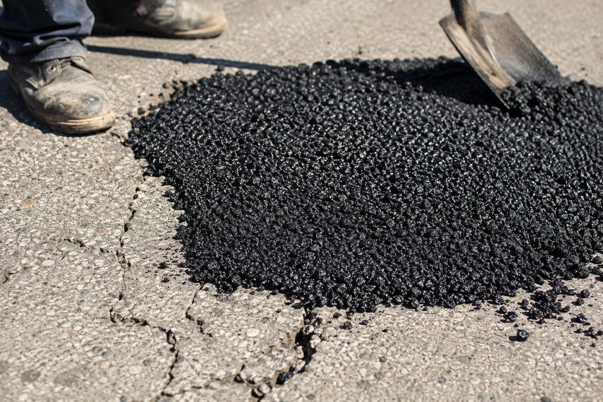 Close-up of black asphalt patch material pressed into a crack in a gray street surface, with a shovel blade and work boots at the edge of the frame in harsh midday light.