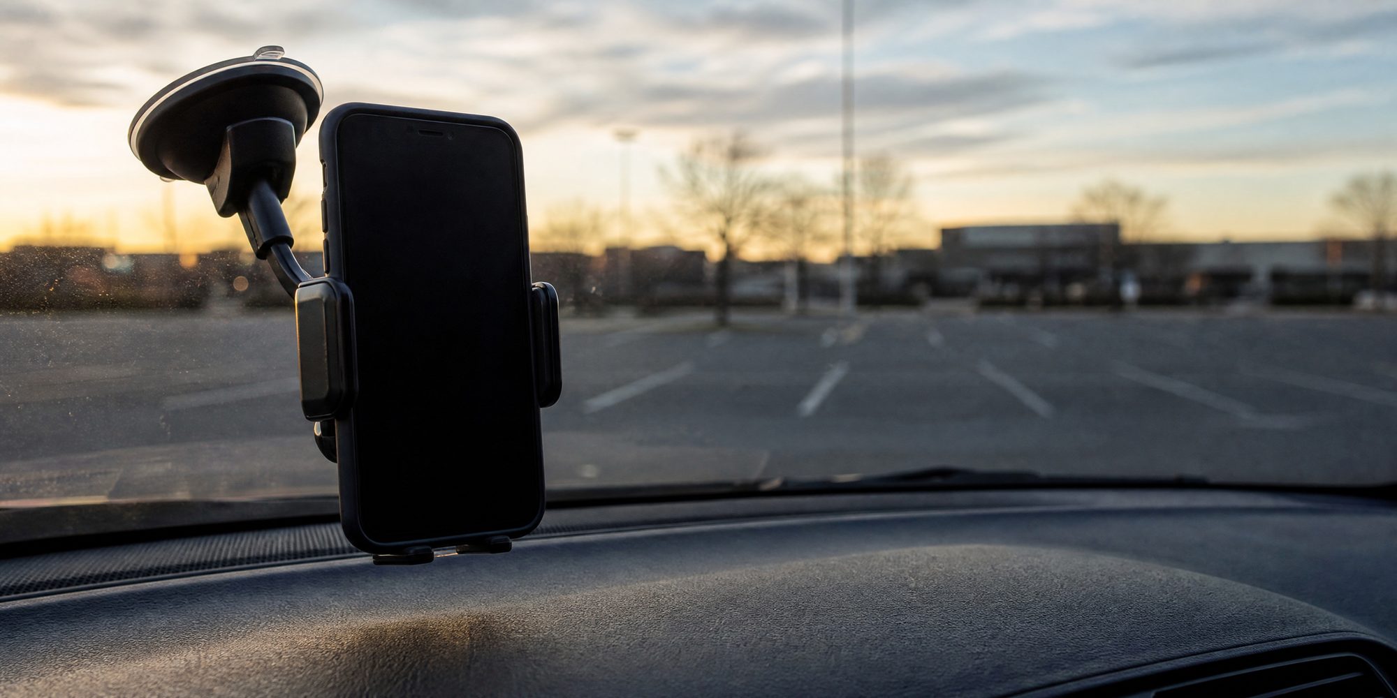 Close macro of a smartphone clamped in a windshield mount inside a parked car, screen dark and unreadable, empty parking lot beyond the glass at first light.