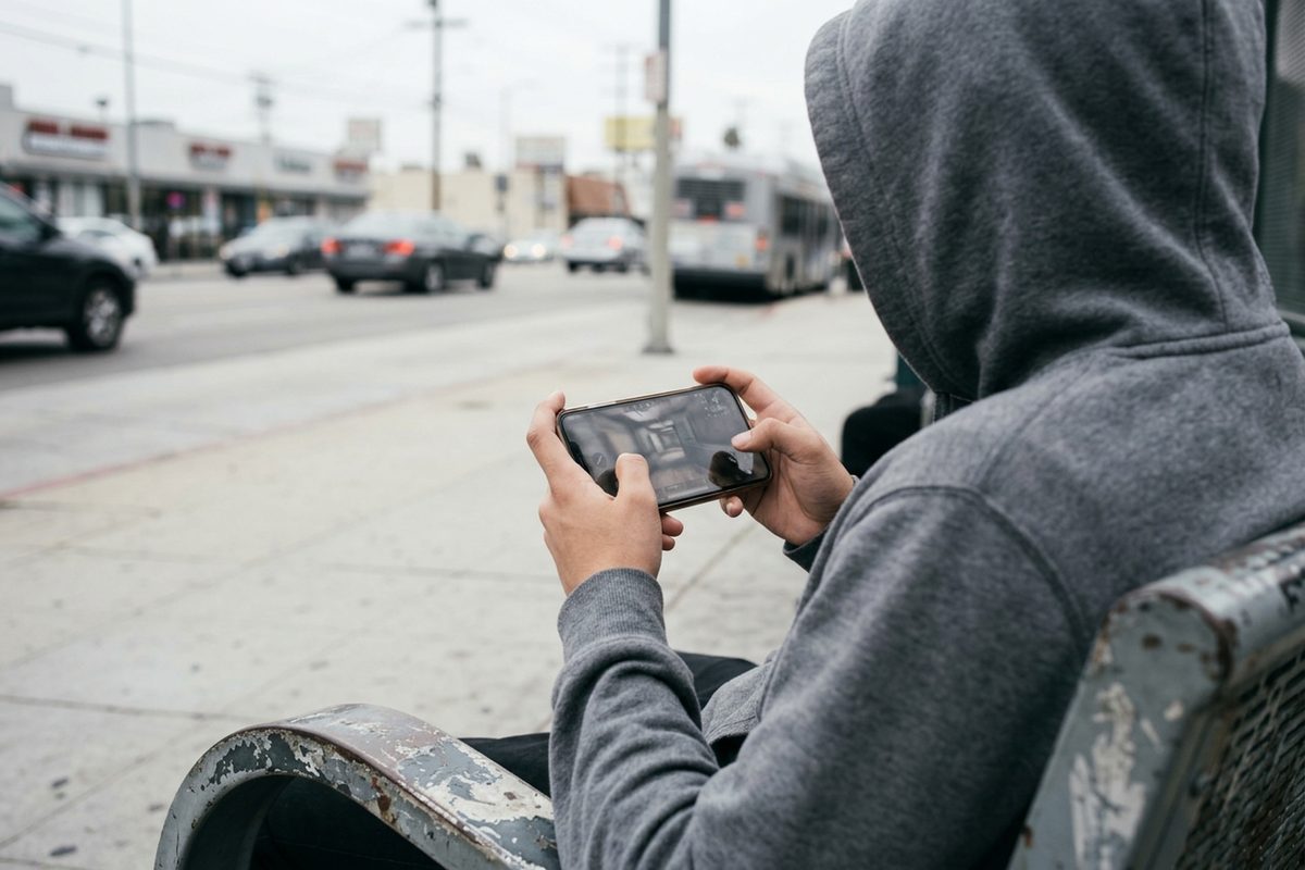 Over-the-shoulder view of a person holding a phone sideways and playing a mobile shooter game at a Los Angeles bus stop on an overcast morning.