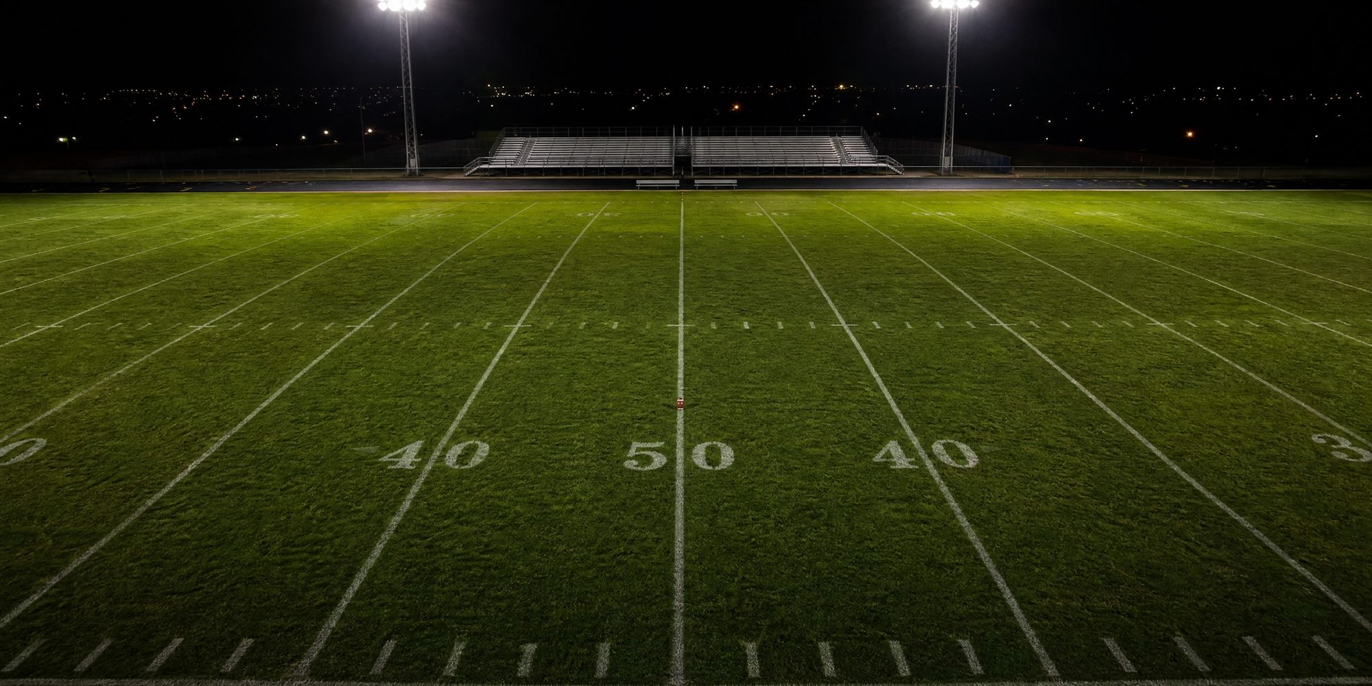 Overhead view of an empty football field at night, chalked yard lines and hash marks lit by stadium lights, with a ball on a tee near midfield.