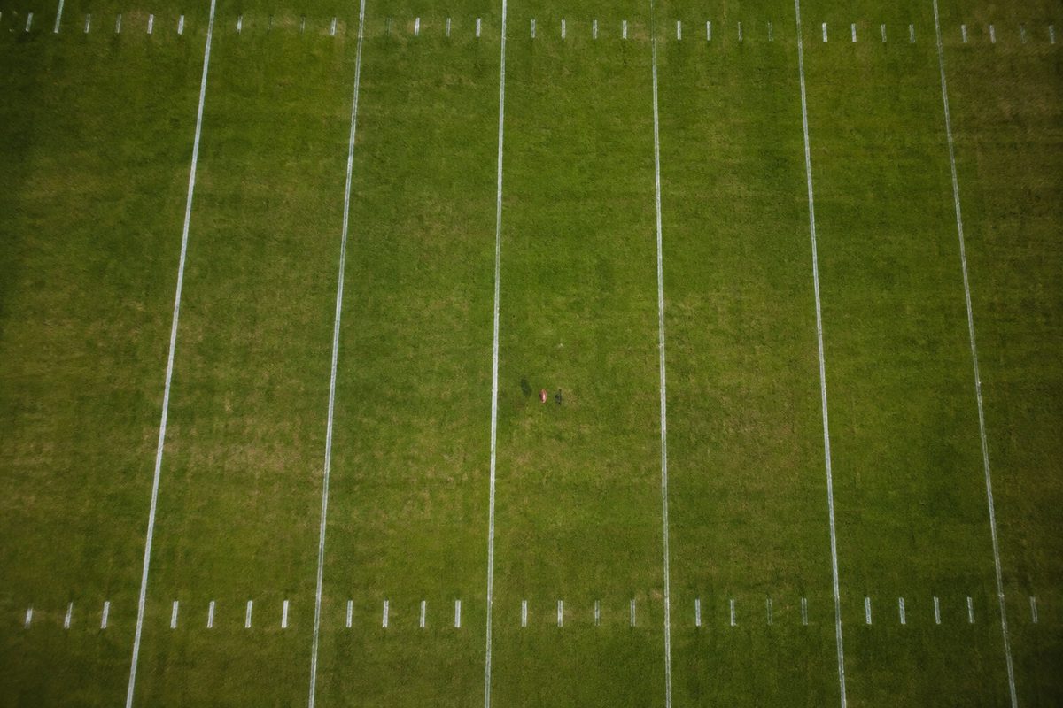 Overhead view of an empty football field at night, chalked yard lines and hash marks lit by stadium lights, with a ball on a tee near midfield.
