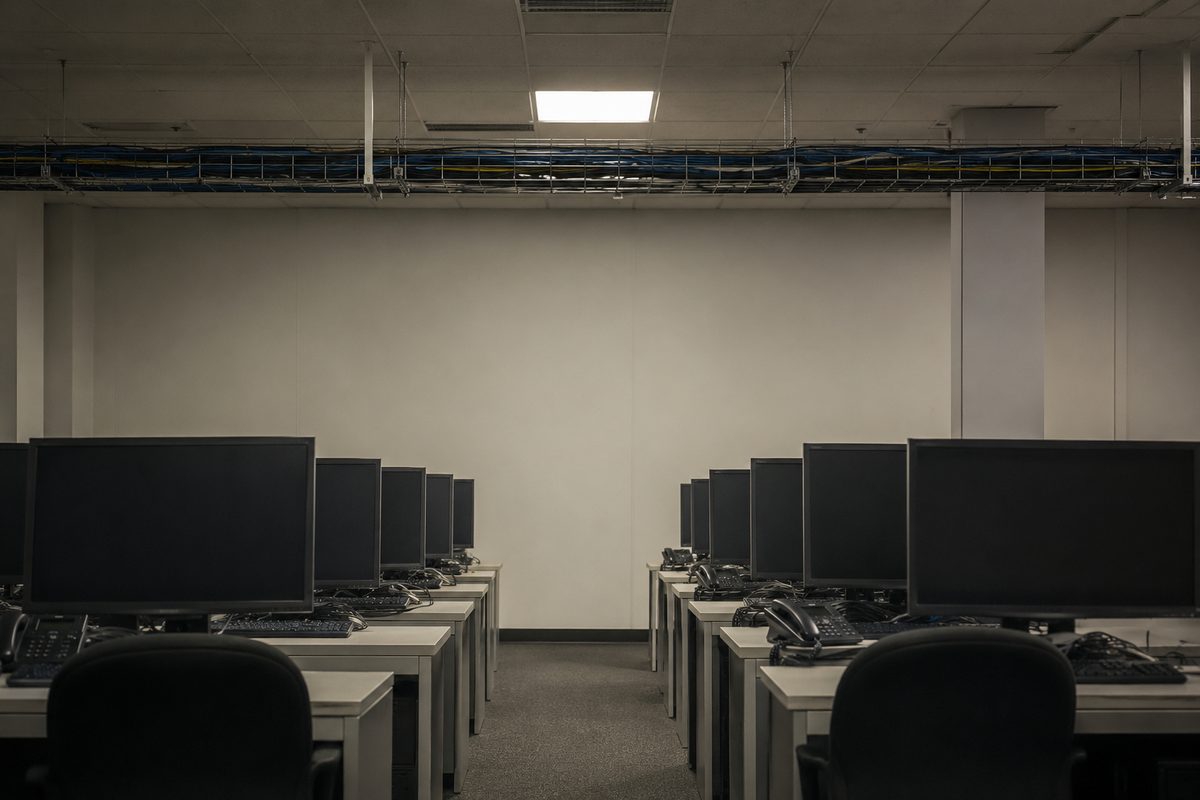 A frontal, symmetrical view of an anonymous back-office room: rows of identical desktop terminals with unreadable screens under flat fluorescent ceiling light.