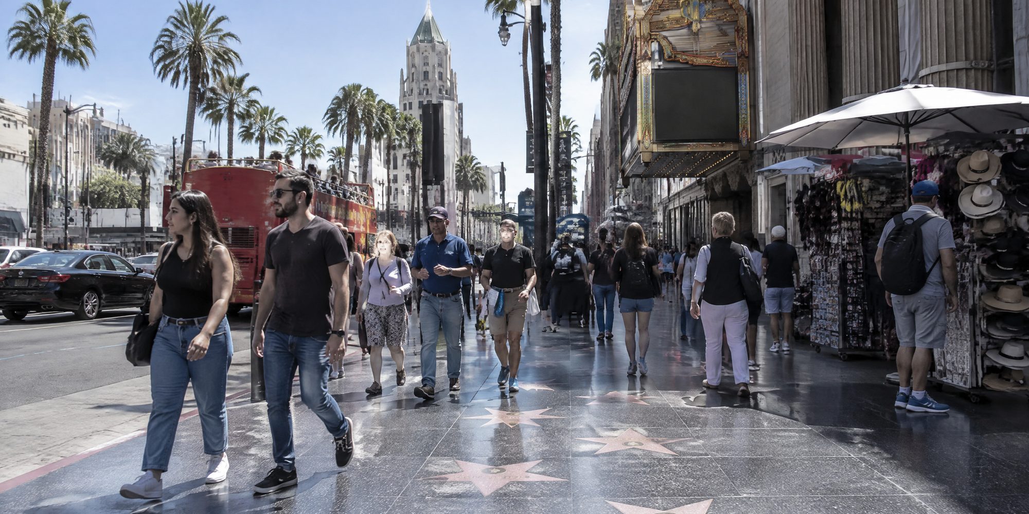 A sunlit Hollywood Boulevard sidewalk inlaid with five-pointed terrazzo and brass stars, people walking across them past a souvenir stand and an open-top sightseeing bus, a theatre marquee and palm trees behind