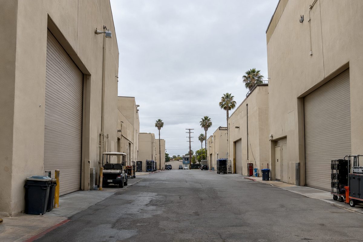 Photo illustration: A quiet lane between rows of tall, windowless beige soundstage buildings on a working film and television studio lot in Los Angeles, seen from ground level at the end of the lane under a flat overcast morning sky. Big rolling stage doors are shut, a parked golf cart and a few equipment carts sit along the curb, and palm trees and power lines show above the rooflines. Nobody is in the frame, and there's no text, signage or logos anywhere in the picture.