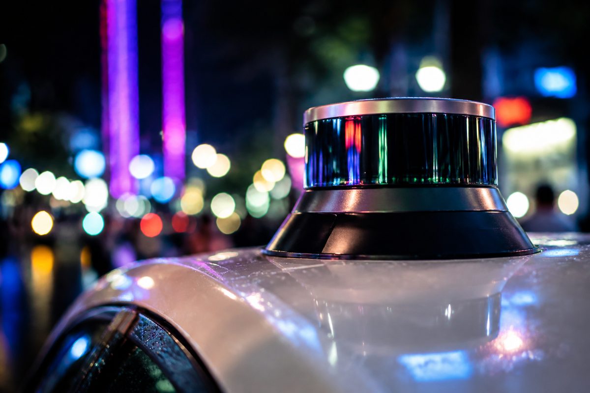Extreme close-up of a roof-mounted spinning laser sensor on a car, unbranded, with colored night lighting reflected in its housing.