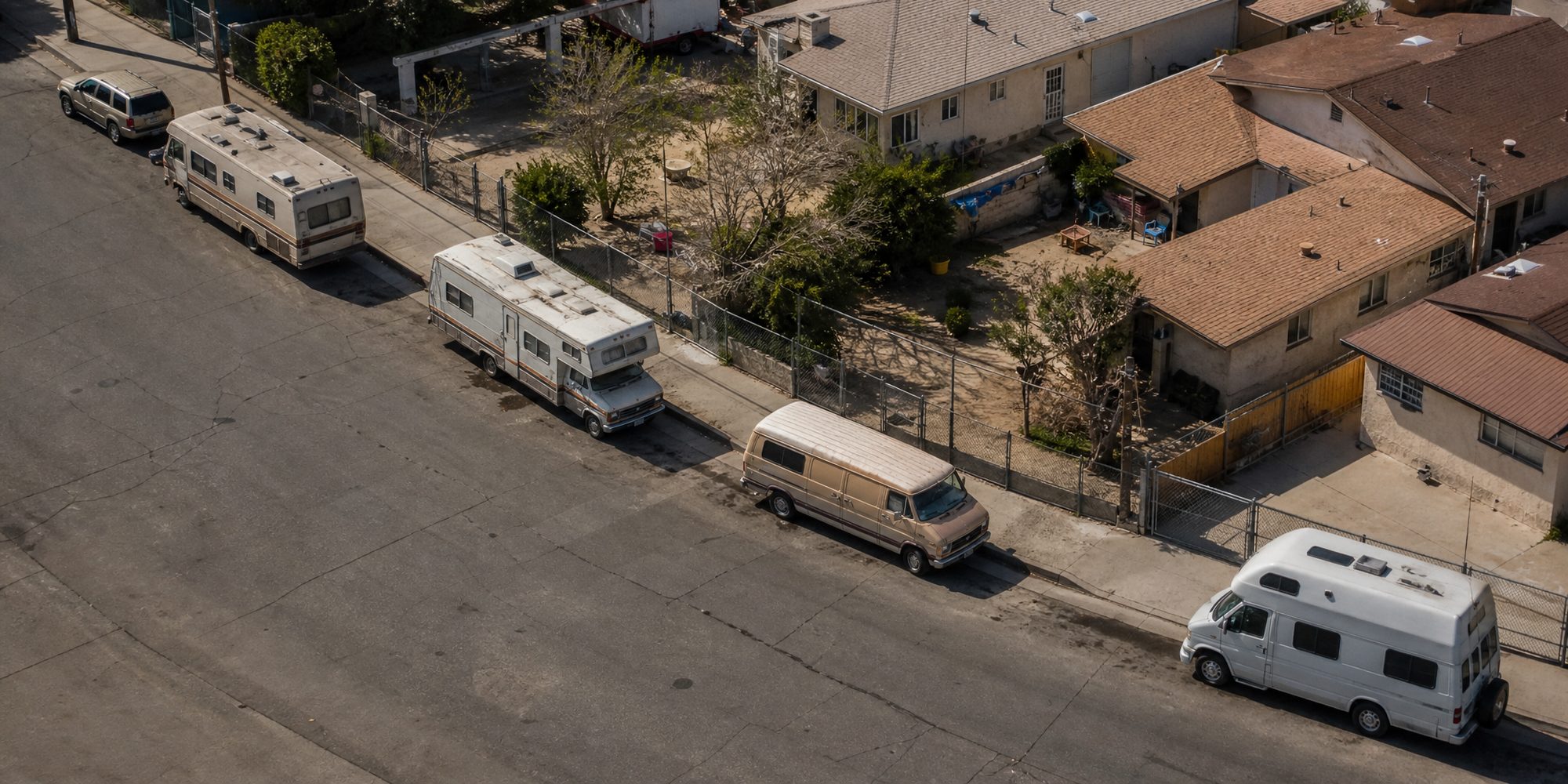 Overhead view of a residential block at midday, with several older RVs and vans parked along one curb beside stucco rooftops and chain-link fencing.