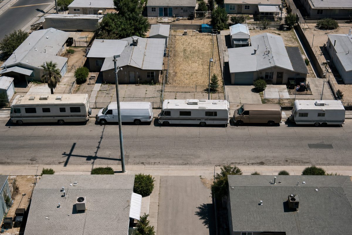 Overhead view of a residential block at midday, with several older RVs and vans parked along one curb beside stucco rooftops and chain-link fencing.