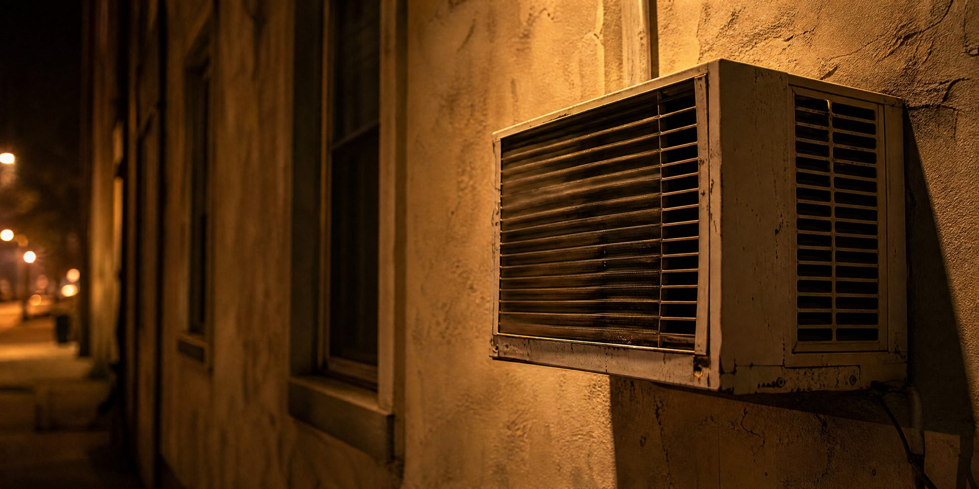 Close-up of a weathered window air-conditioning unit mounted in a stucco wall, grille vibrating, lit by orange evening street light.