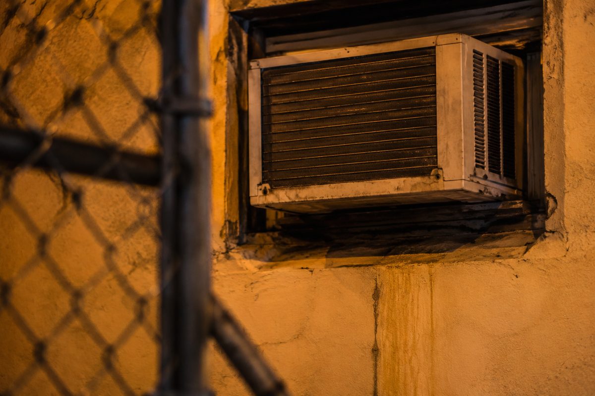 Close-up of a weathered window air-conditioning unit mounted in a stucco wall, grille vibrating, lit by orange evening street light.