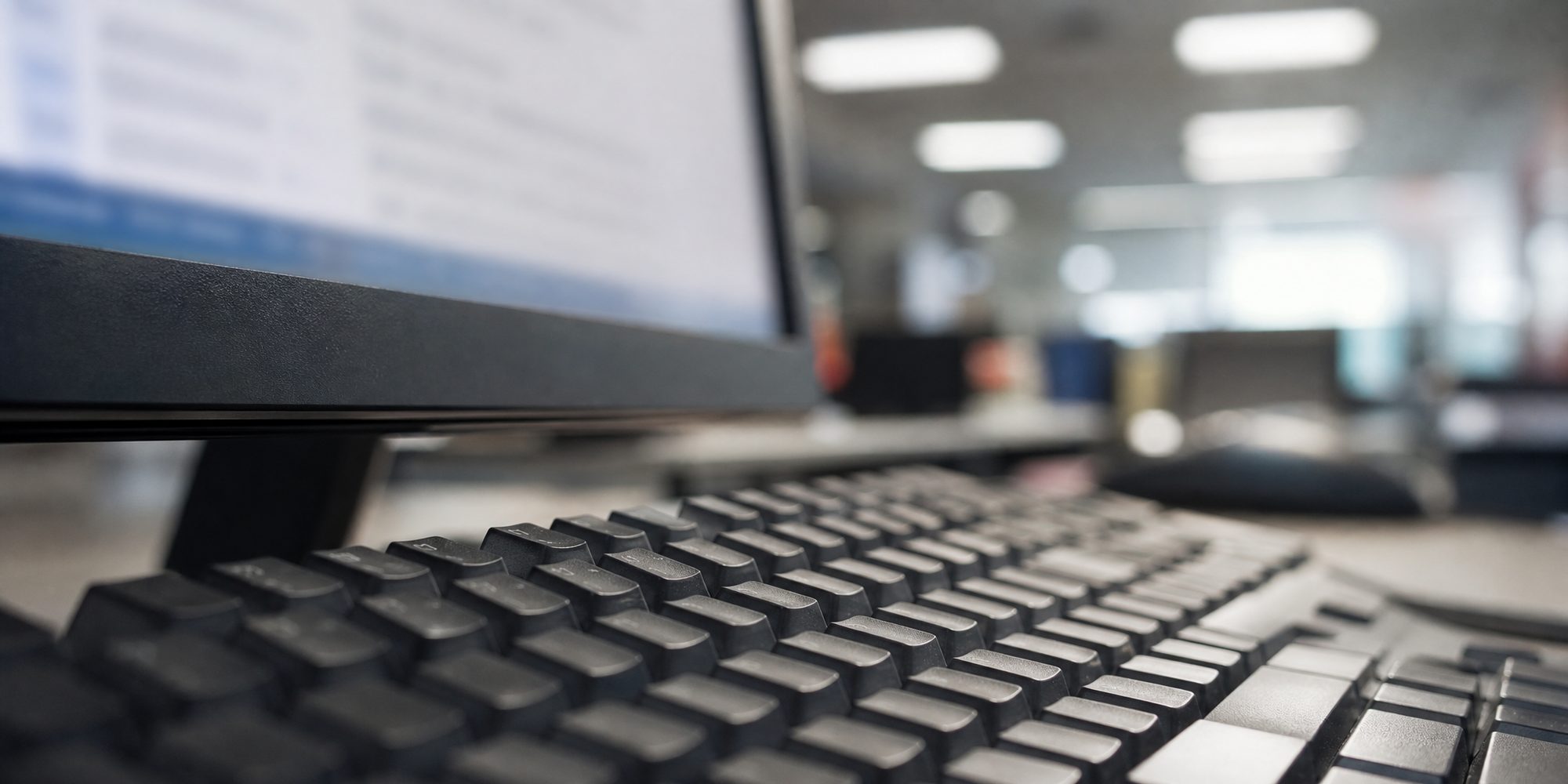 Close macro of an unbranded keyboard and the edge of a monitor showing blurred, unreadable text under fluorescent office light.