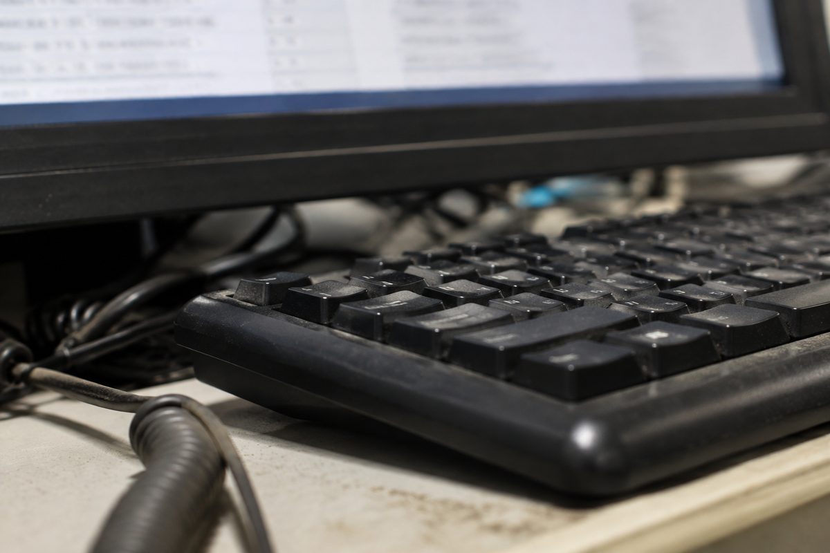 Close macro of an unbranded keyboard and the edge of a monitor showing blurred, unreadable text under fluorescent office light.
