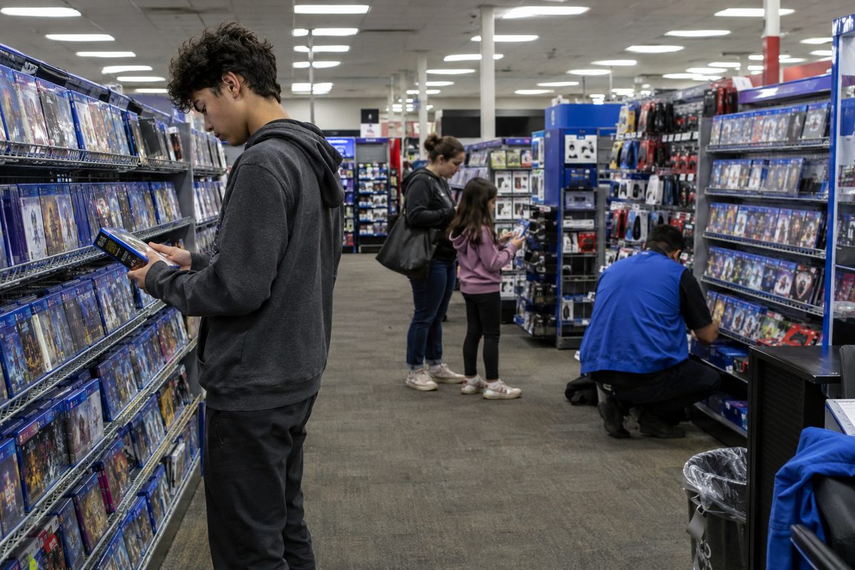 Shoppers browse shelves of video games under fluorescent lights in a Burbank electronics store, one holding a game case.