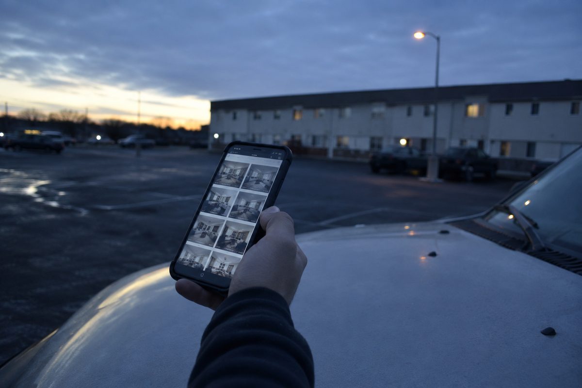 Close view of a hand holding a phone at dawn, the screen showing an unreadable grid of apartment listings, with a low stucco apartment block out of focus behind an empty parking lot.