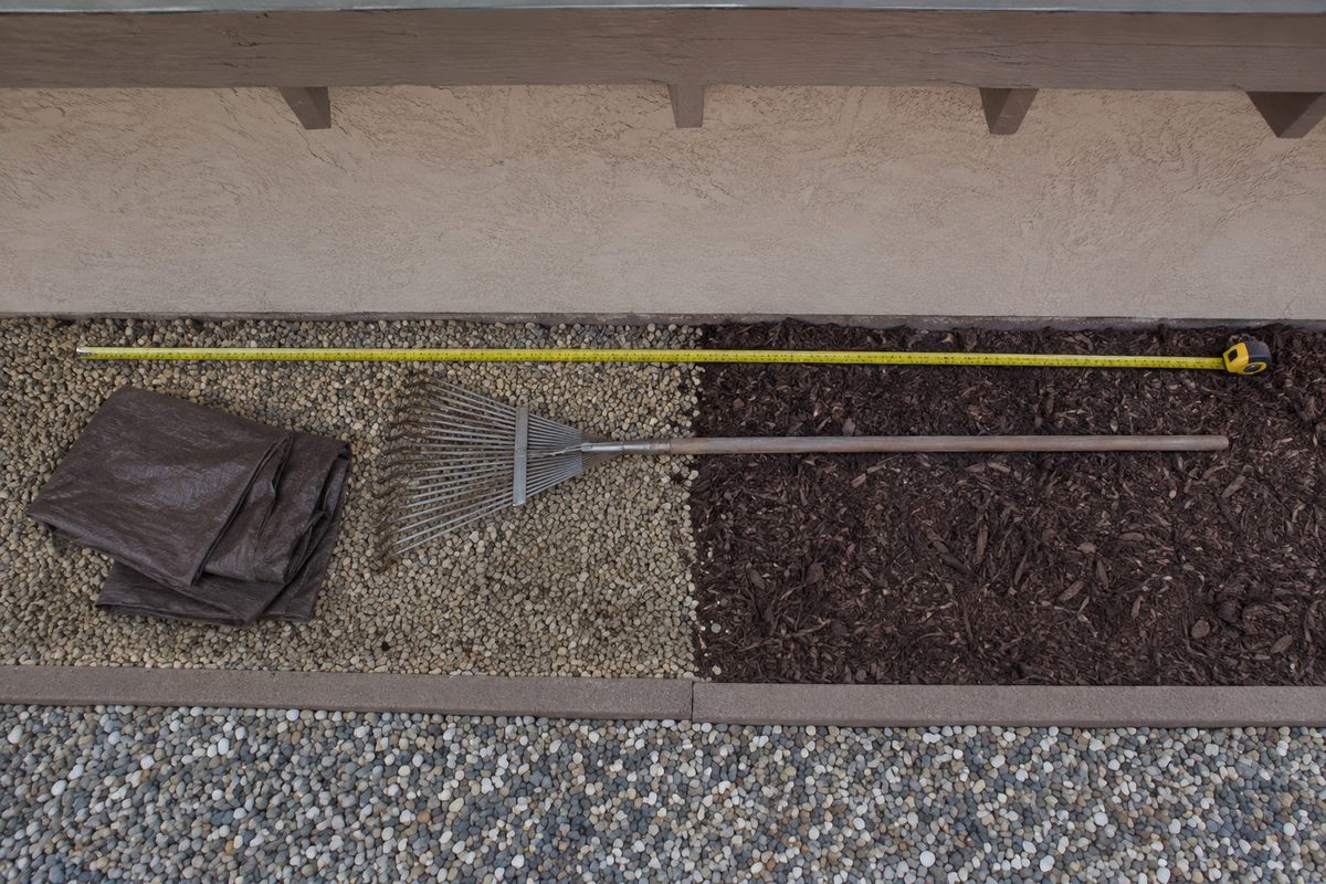 Photo illustration: overhead flat-lay of a five-foot strip of ground against a stucco wall under a shallow eave — a tape measure pulled out to five feet, a folded tarp, a rake, gravel on one side and bark mulch on the other, neutral daylight, no house identifiable, no people.