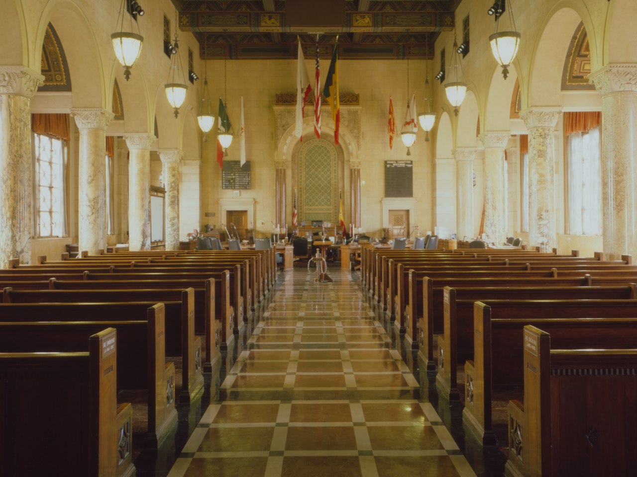 The Los Angeles City Council chamber seen from the back: empty public benches down a checkerboard aisle, the members' desks and flags at the far end