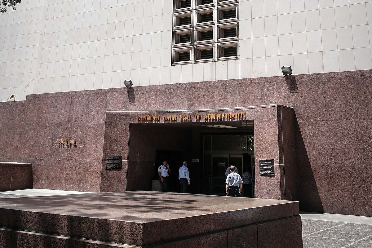 The entrance of the Kenneth Hahn Hall of Administration in downtown Los Angeles, the building's name in gold lettering above the doorway, three people walking in past the granite plaza