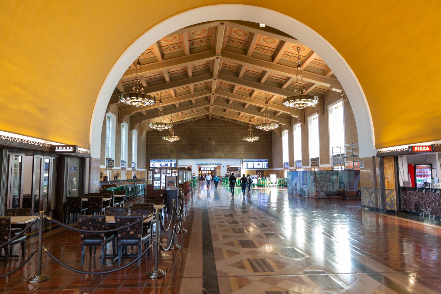 The main concourse of Los Angeles Union Station, a barrel-vaulted hall with hanging chandeliers and a polished terrazzo floor, a few travelers walking toward the arrivals and departures boards at the far end