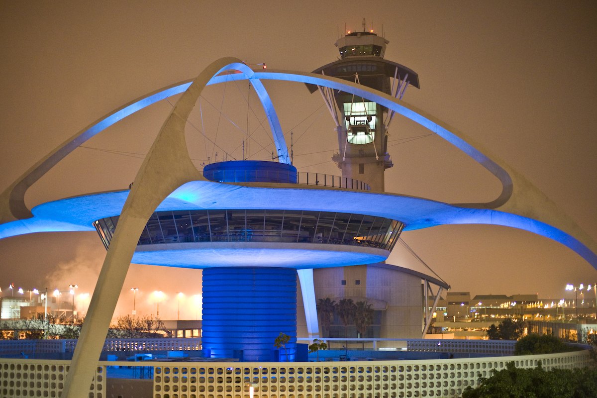 The Theme Building at Los Angeles International Airport at night, its saucer-shaped top lit blue beneath a white curving arch, with the airport control tower rising behind it and lights glowing across the airfield.
