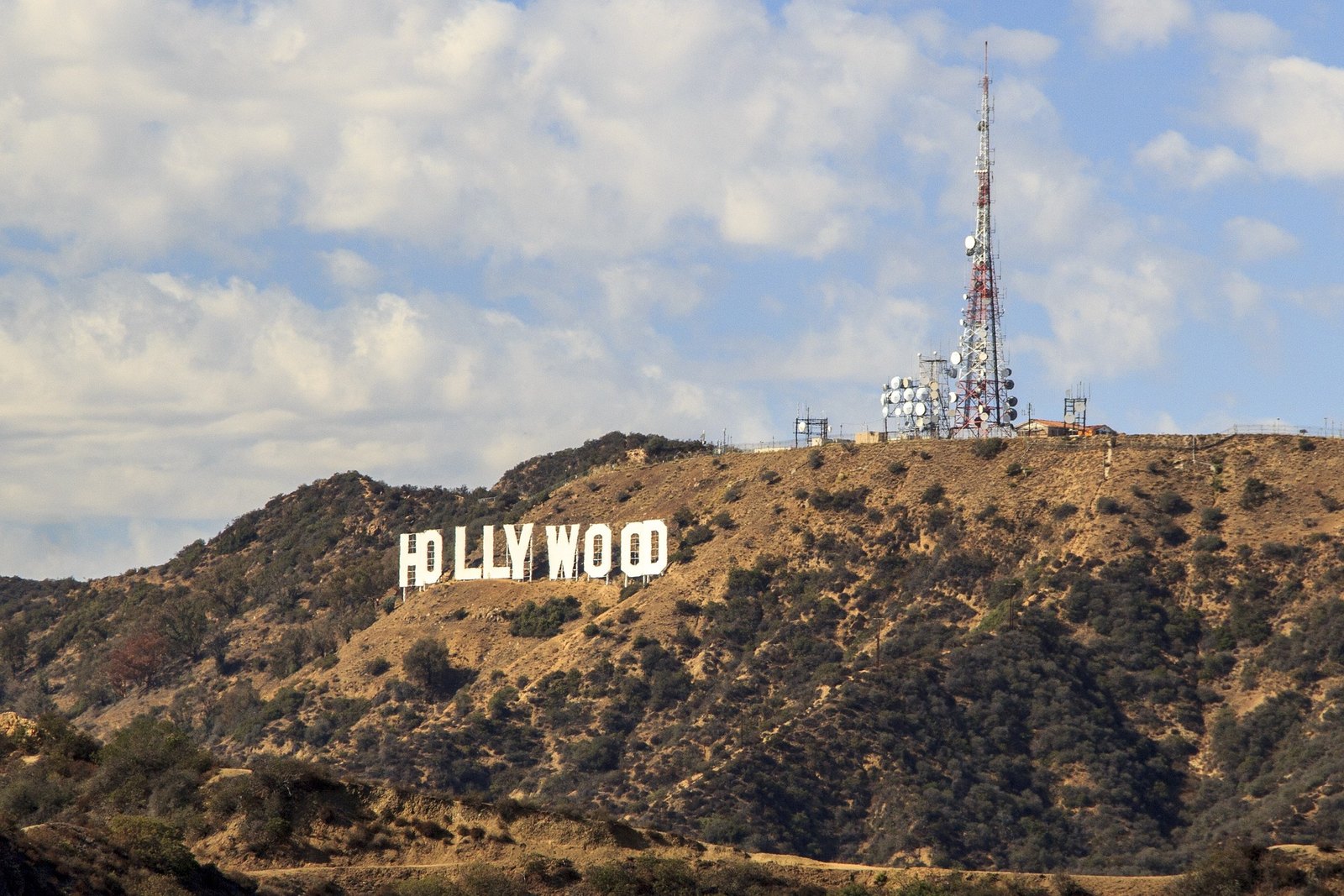The white Hollywood letters sit on a dry brown hillside under a partly cloudy blue sky, with a tall red and white antenna tower and satellite dishes on the ridge beside them.