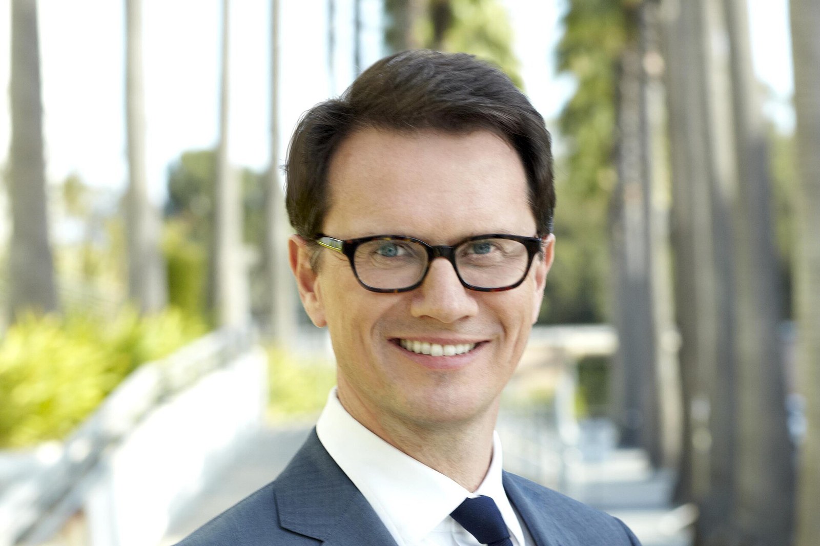 Peter Rice, smiling, in glasses and a gray suit, on a palm-lined walkway in Los Angeles