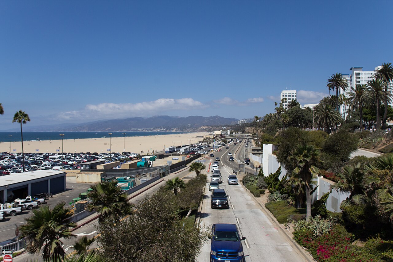 Moomat Ahiko Way descending from Ocean Avenue toward Pacific Coast Highway under the Santa Monica Pier, with the beach parking lots and the ocean to the west and the highway running north toward the bluffs