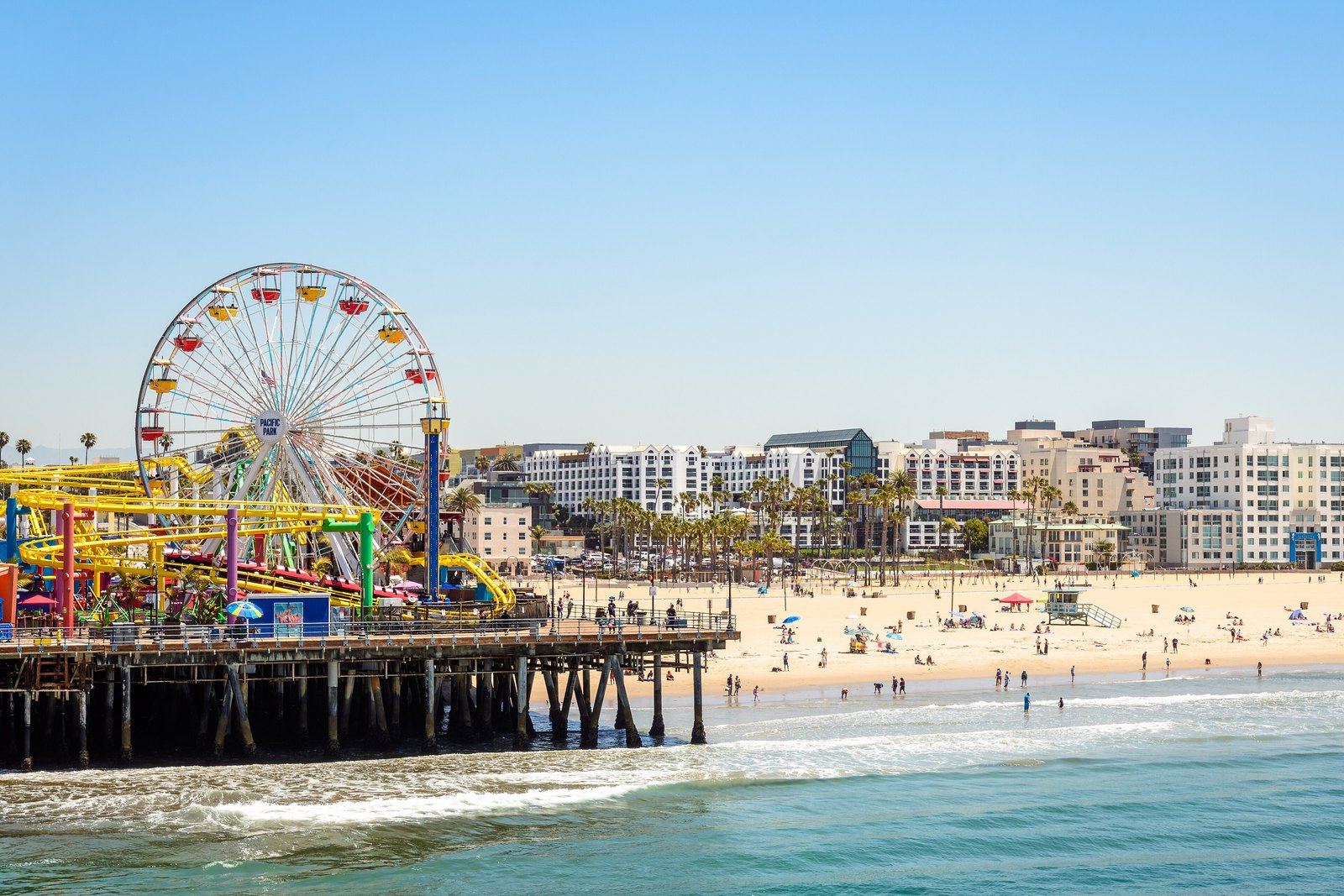 Pacific Park's colorful Ferris wheel and roller coaster track on the Santa Monica Pier, with a sandy beach, white hotels and palm trees behind, and people on the sand and in the ocean under a clear blue sky.