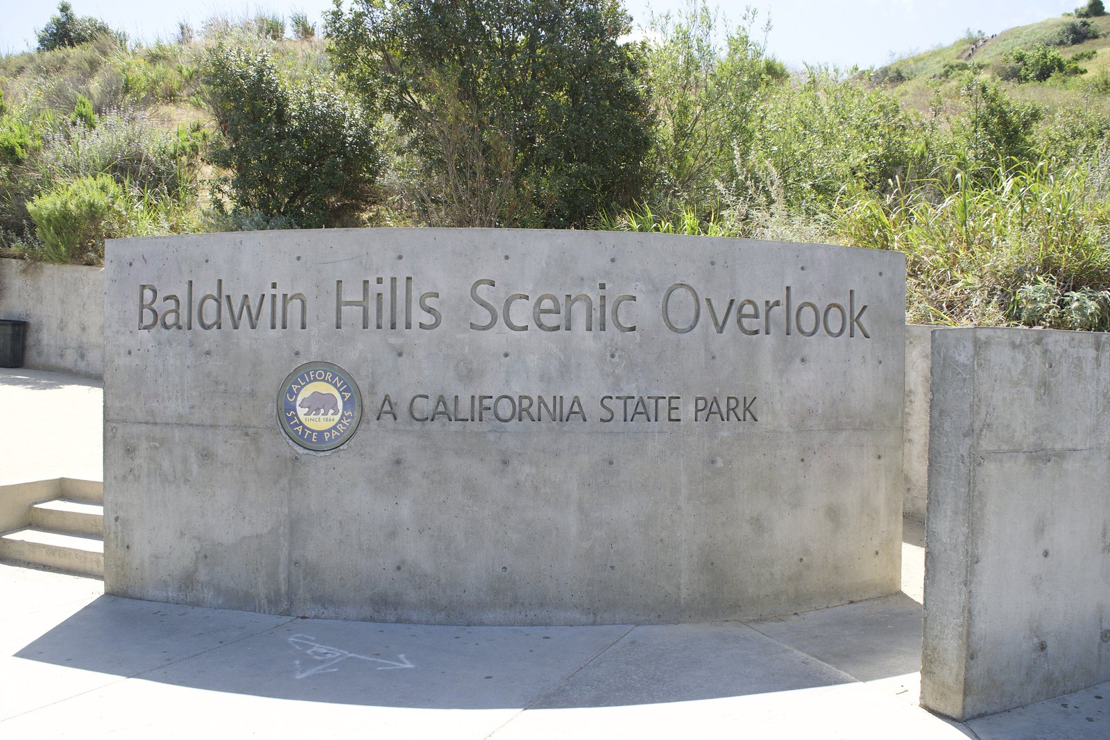 Curved concrete sign reading Baldwin Hills Scenic Overlook, with a California State Parks seal and the words A California State Park, in front of green hillside shrubs under a bright sky.