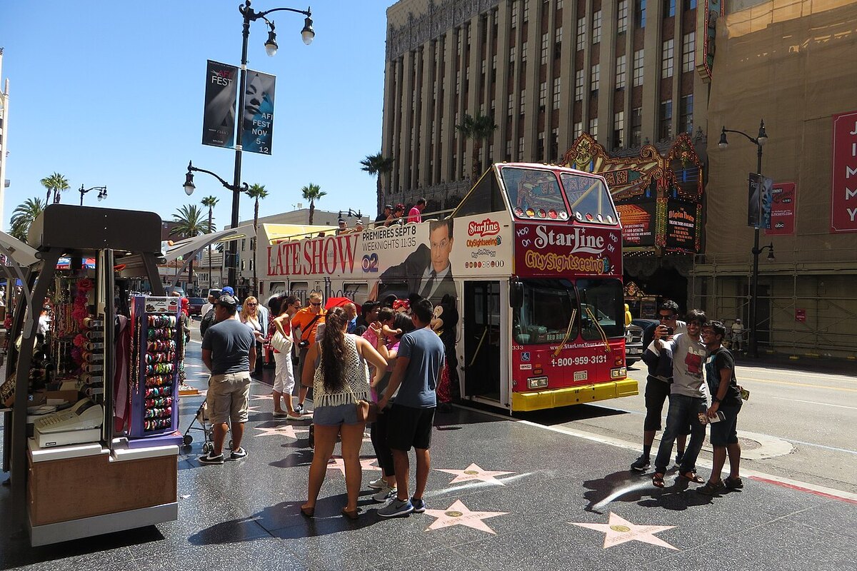 A sunlit Hollywood Boulevard sidewalk inlaid with five-pointed terrazzo and brass stars, people walking across them past a souvenir stand and an open-top sightseeing bus, a theatre marquee and palm trees behind
